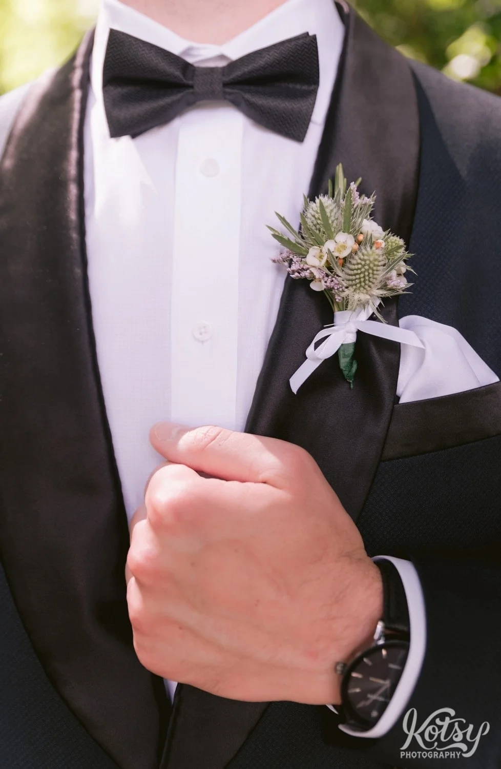 A close up shot of a groom's hand holding on to the lapel of a black tuxedo. A black bowtie and boutonniere is also seen
