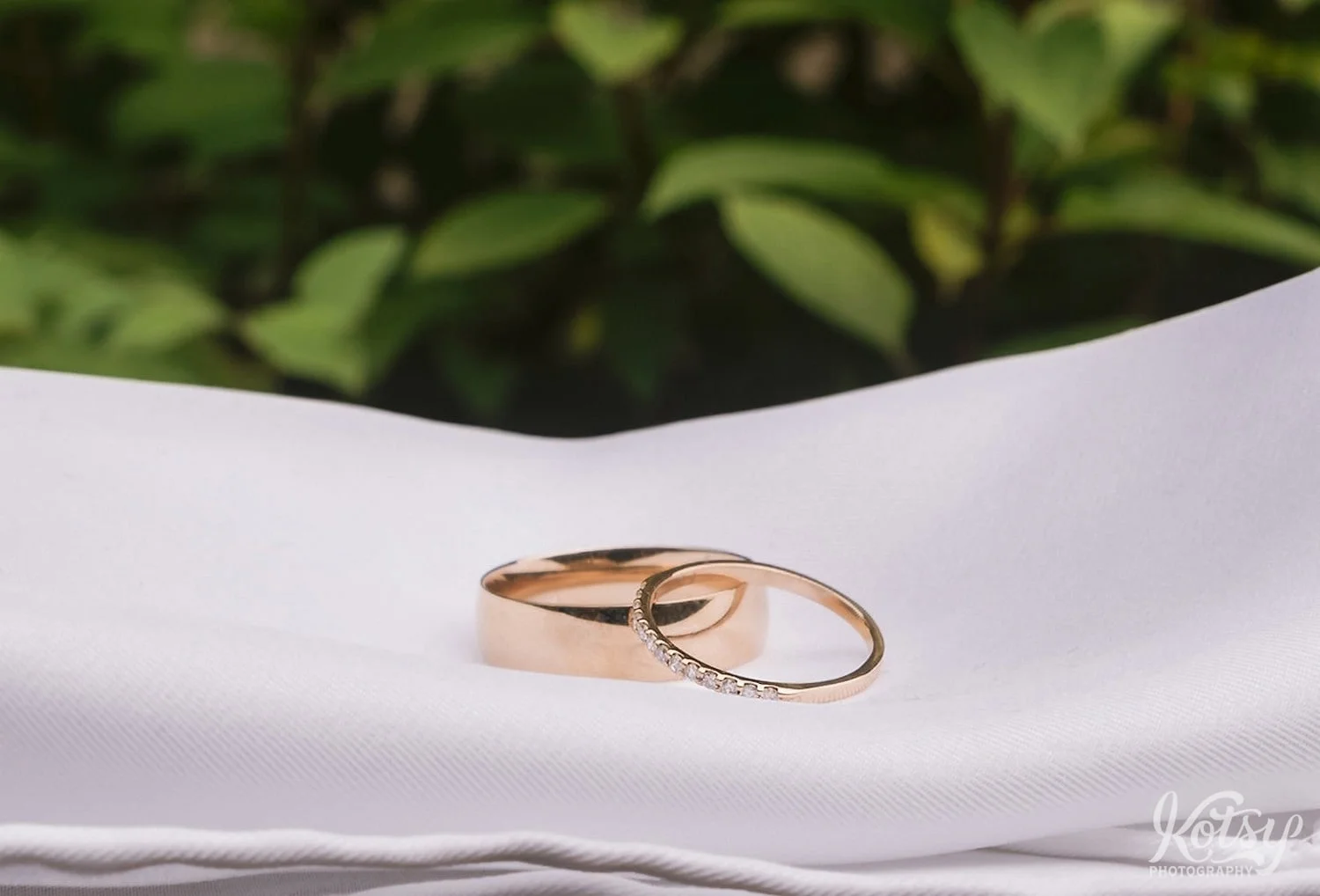 Two wedding bands are seen on a white cloth with green plants in the background