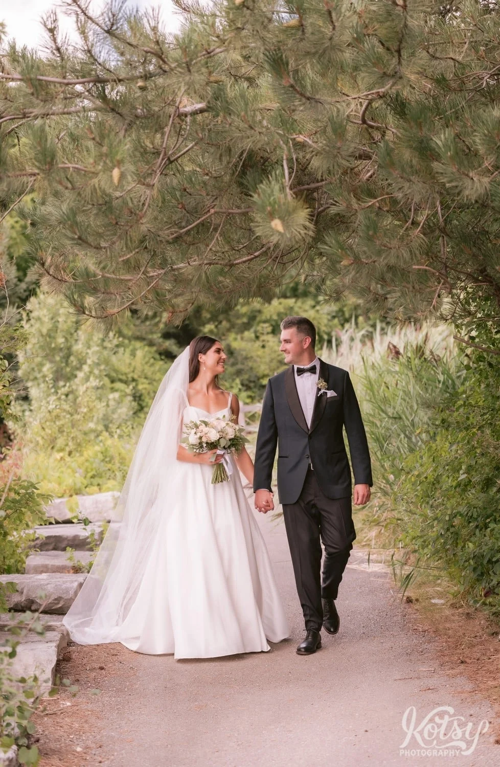 A wide shot of a bride and groom walk on a gravel path while smiling at each other. Photographed at Hunter's Point Wildlife Park in Toronto, Ontario, Canada
