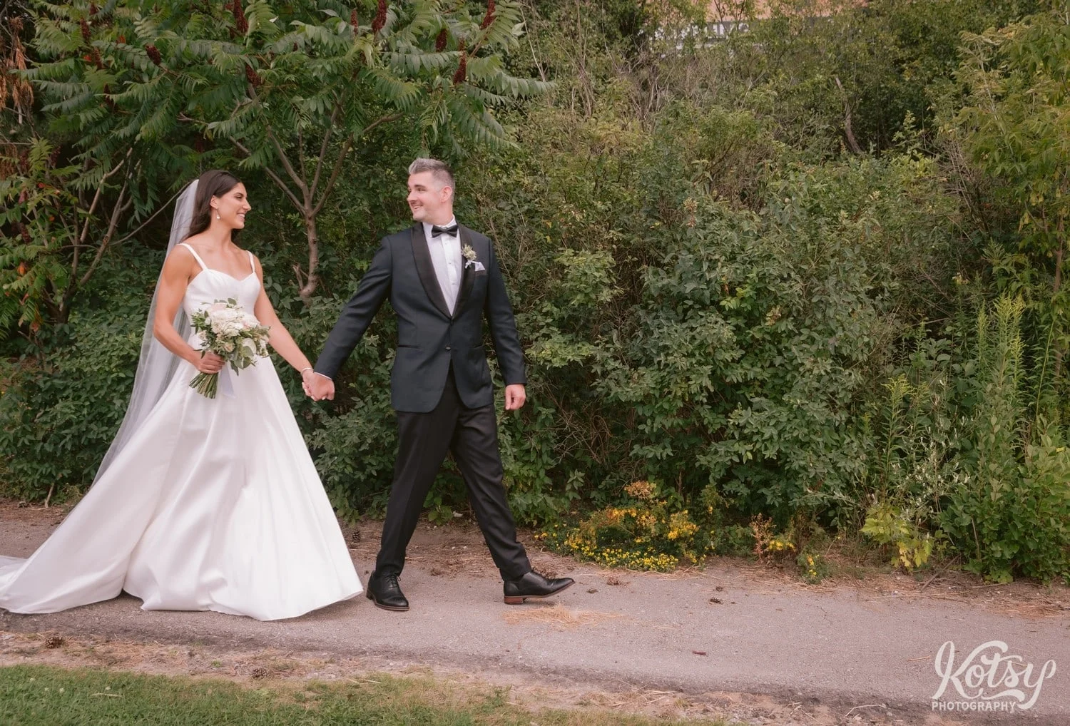 A wide shot of a groom walking with his bride while holding her hand. Photographed at Hunter's Point Wildlife Park in Toronto, Ontario, Canada
