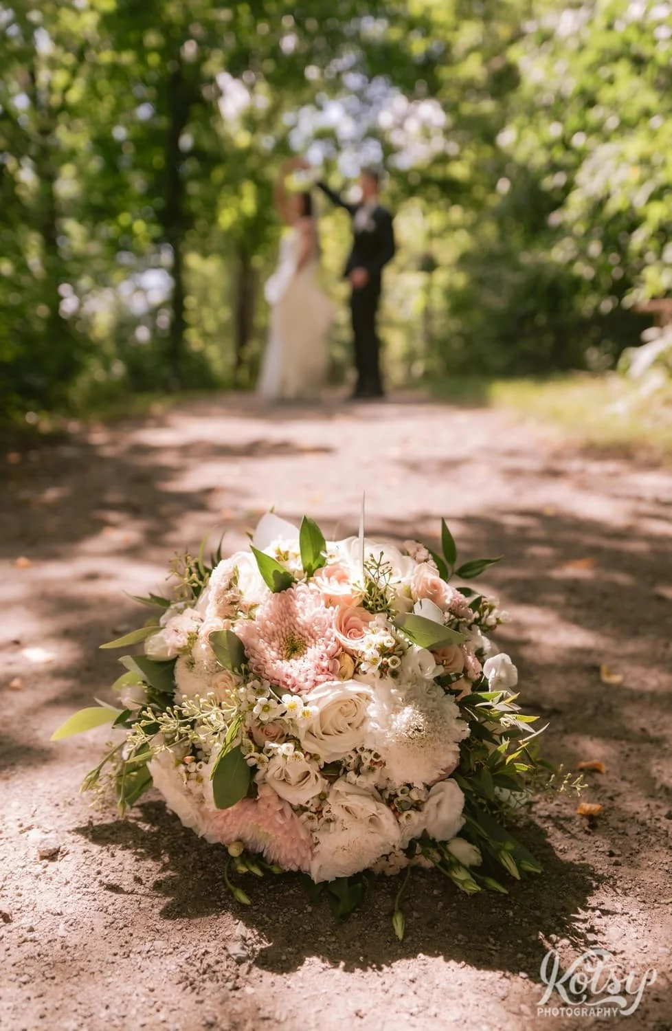 A close up shot of a flower bouquet on a dirt path with an out of focus groom spinning his bride in the background. Photographed at Hunter's Point Wildlife Park in Toronto, Ontario, Canada