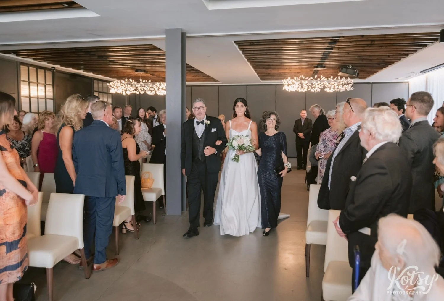 A bride in a white wedding gown walks down an aisle with her parents during a packed house at a Village Loft wedding ceremony in Toronto, Canada