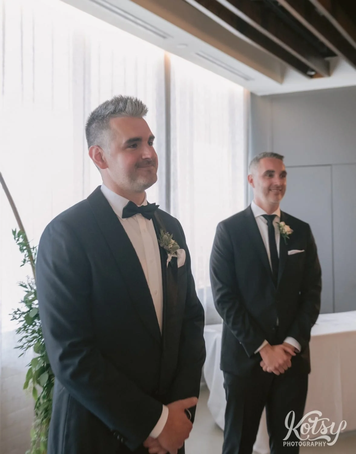 A groom is smiling as he watches his bride walk down the aisle during his Village Loft wedding ceremony in Toronto, Canada