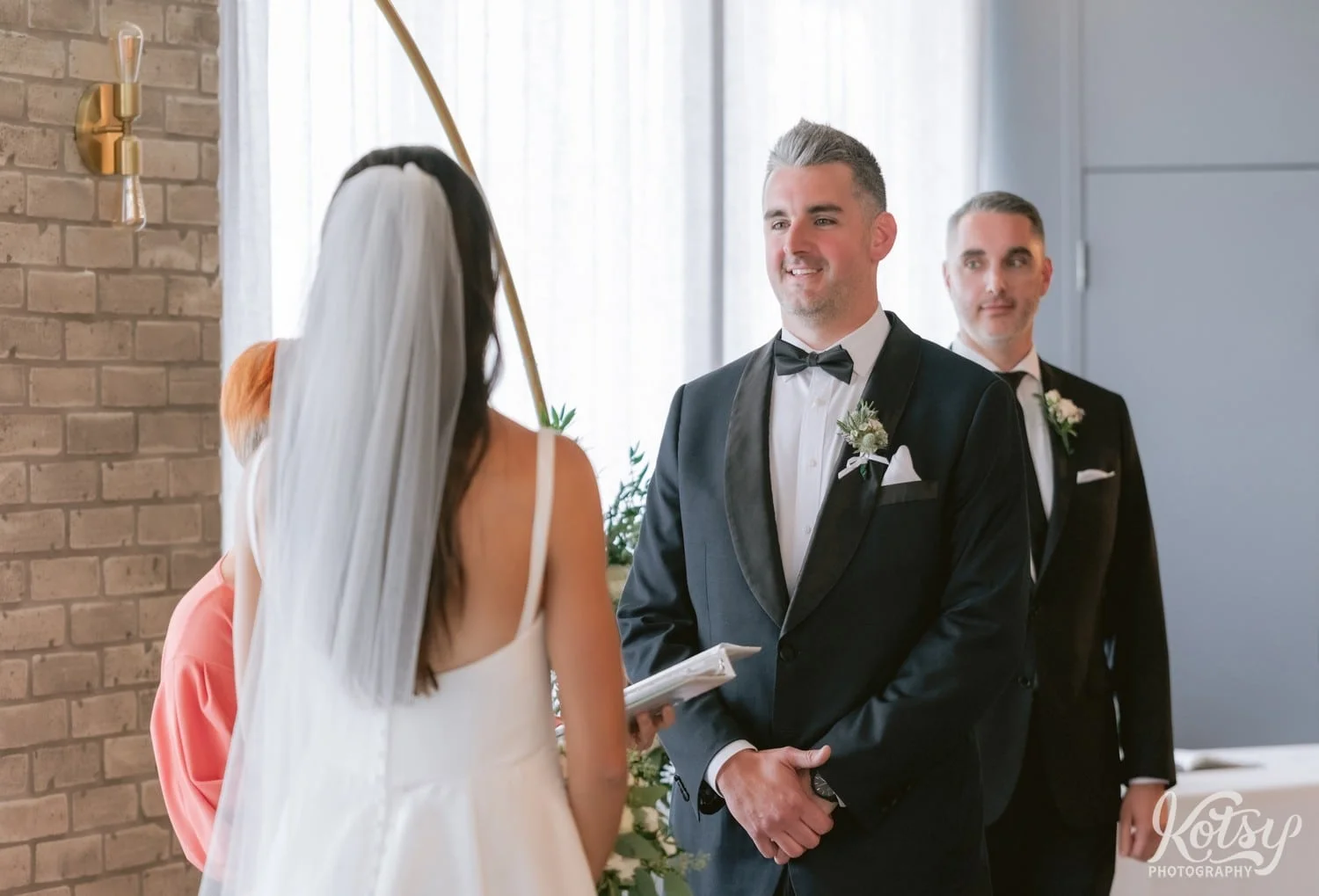 A groom in a black tux smiles as he looks at his bride during their Village Loft wedding ceremony in Toronto, Canada