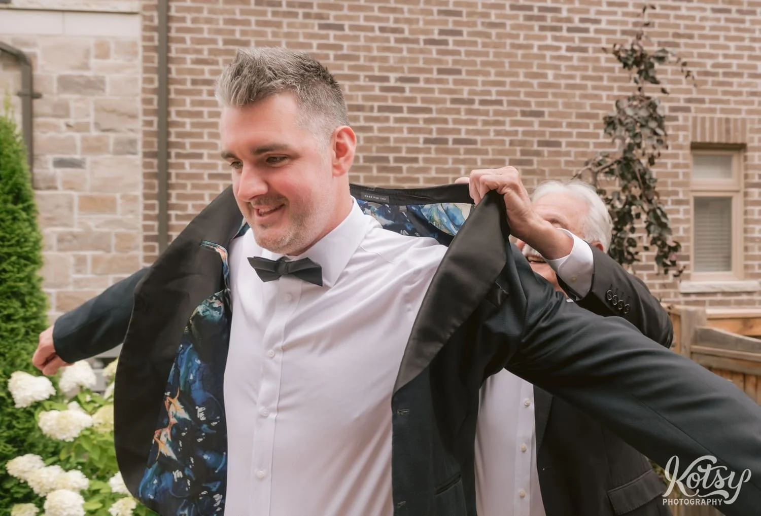 A groom wearing a black bow-tie puts on a black jacket with the help of his father.