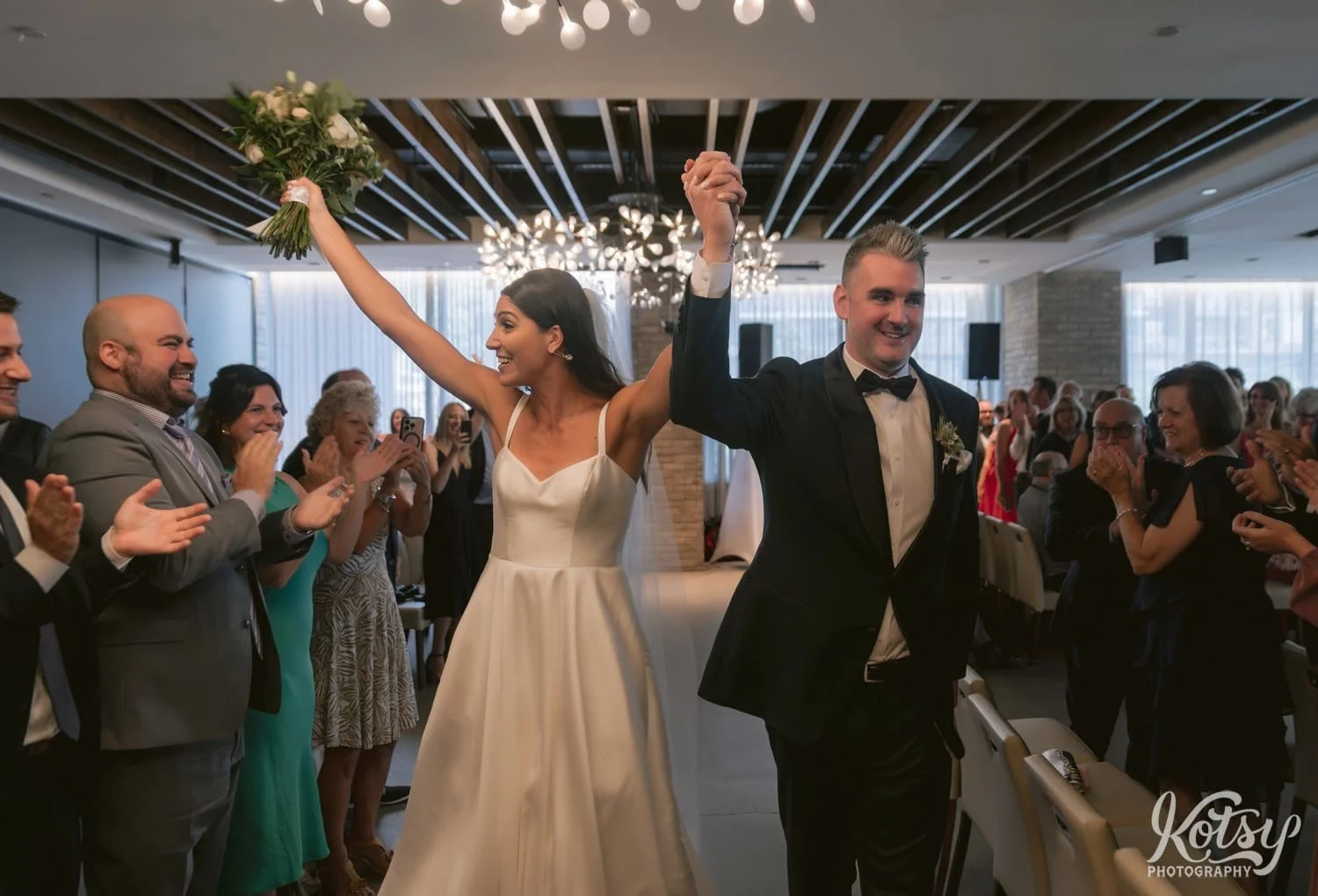 A bride and groom raise their hands in celebration as they walk back up the aisle at the end of their Village Loft wedding ceremonyin Toronto, Canada