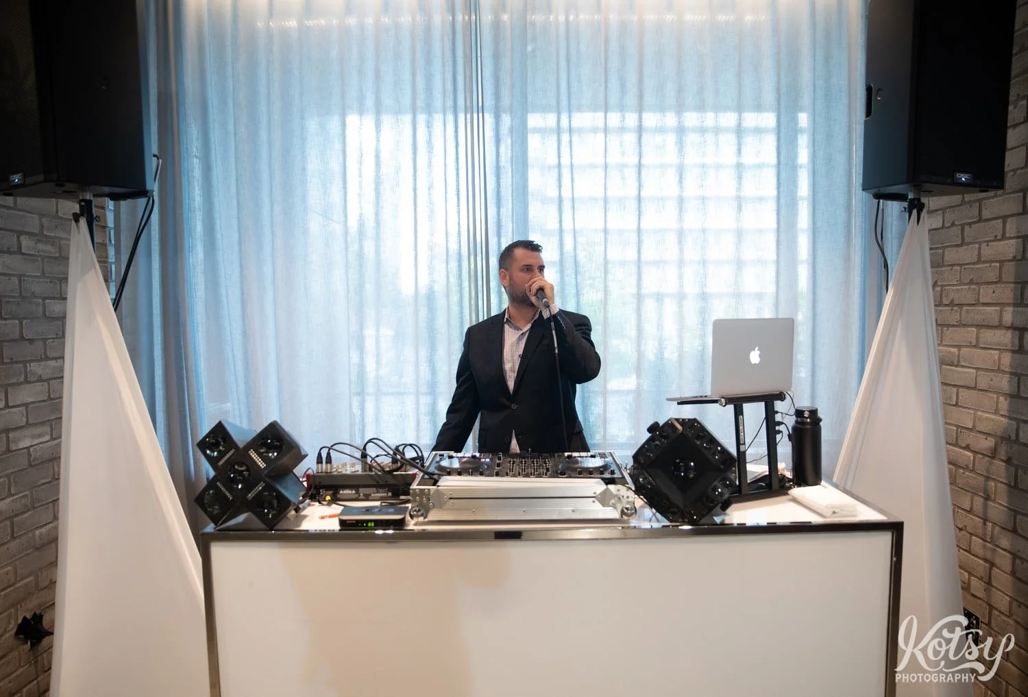 A DJ dressed in a black suit talks into a microphone during a Village Loft wedding reception in Toronto, Canada