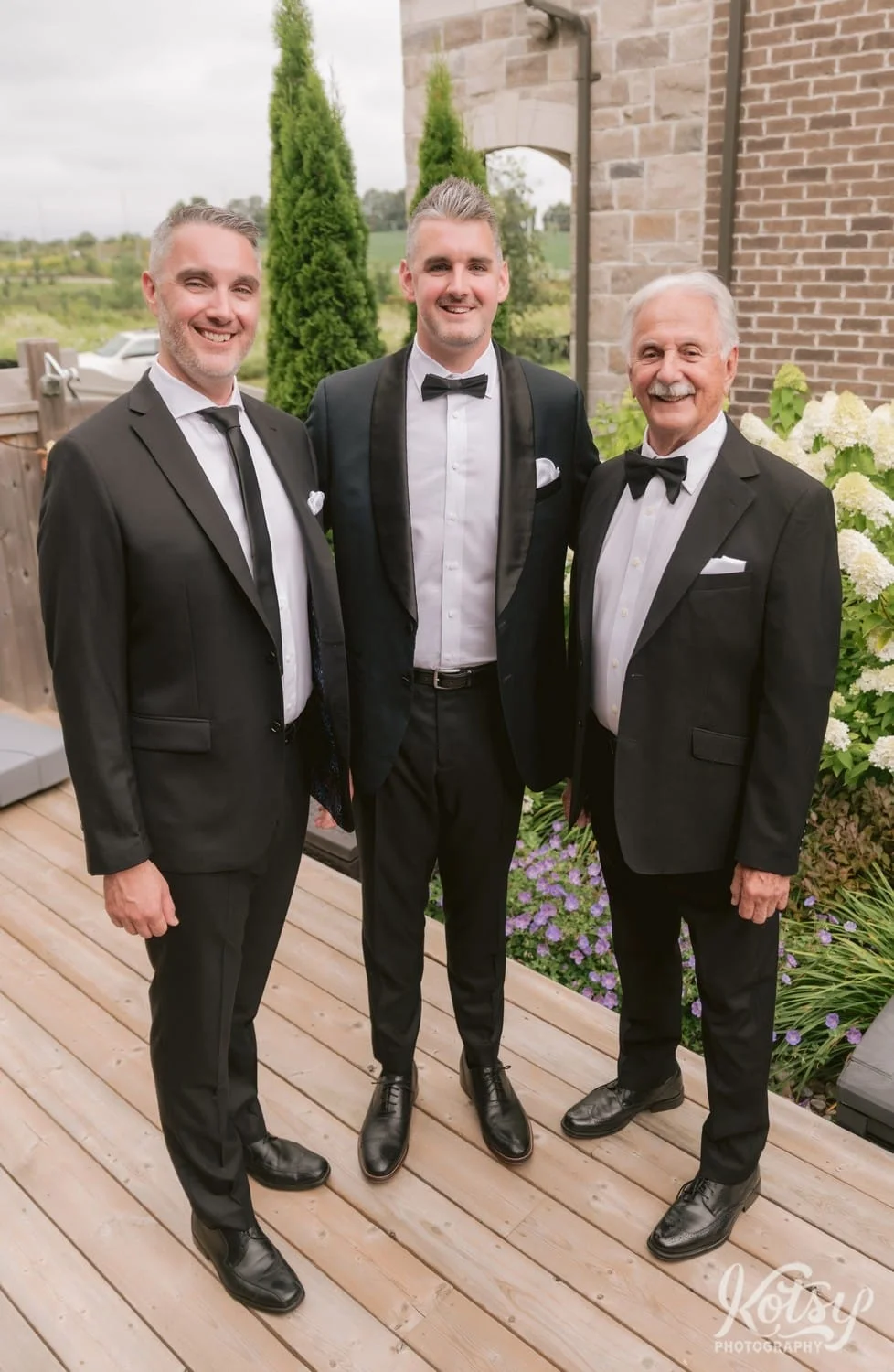 Three men in black suits pose for a group photo on a back patio