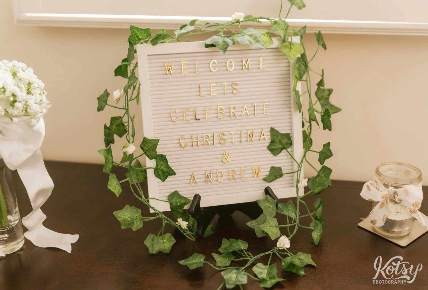 A sign reading "Welcome. Let's Celebrate Christina & Andrew" ss seen on a table with green plant wrapped around the frame
