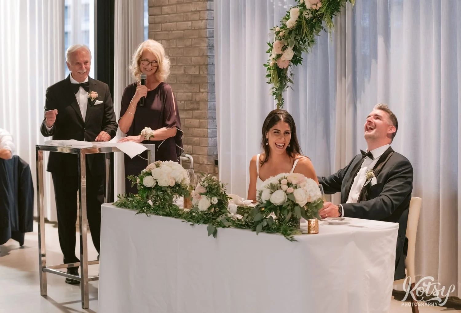 A bride and groom let out a big laugh while the groom's mother makes a speech during their Village Loft wedding reception in Toronto, Canada