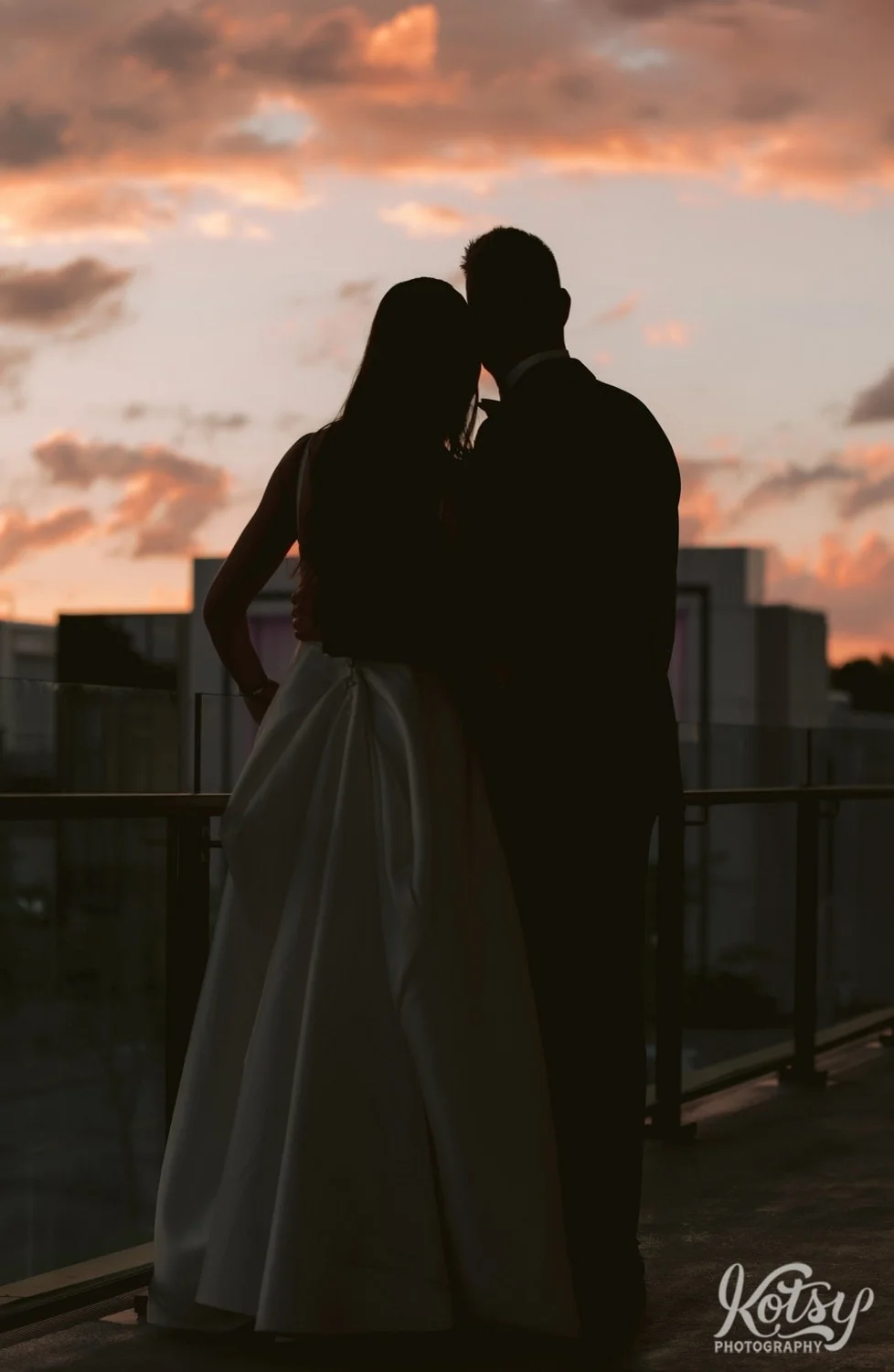 A silhouette of a bride and groom on a balcony with a beautiful sunset ahead of them. Photographed during a Village Loft wedding reception in Toronto, Canada
