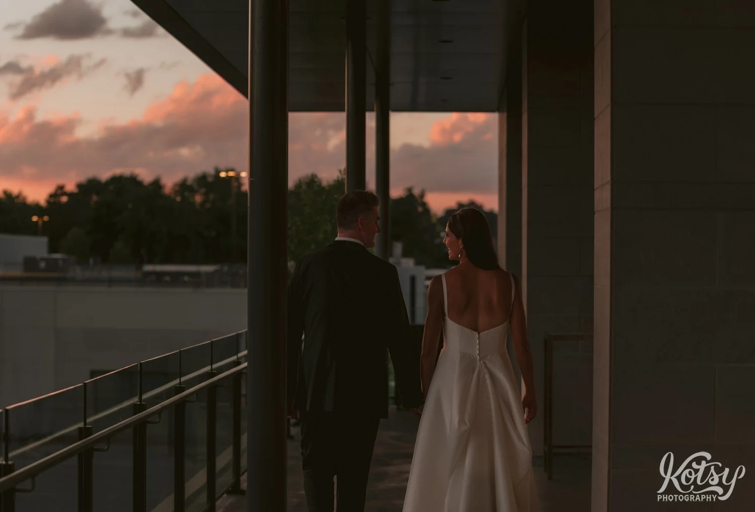 A bride and groom walk away from the camera on a balcony during sunset at their Village Loft wedding reception in Toronto, Canada