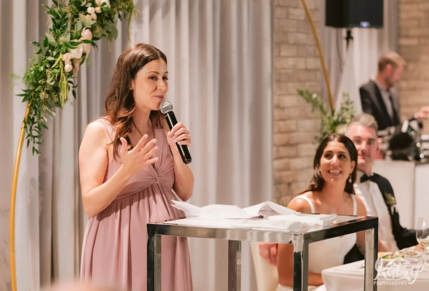 A woman in a pink dress makes a speech while the bride and groom are seen smiling in the background. Photographed during a Village Loft wedding reception in Toronto, Canada