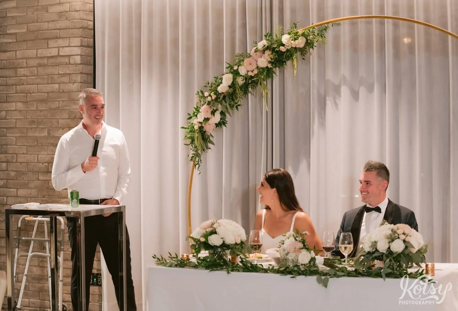 A man in a white shirt smiles as he makes a speech to a bride and groom during their Village Loft wedding reception in Toronto, Canada