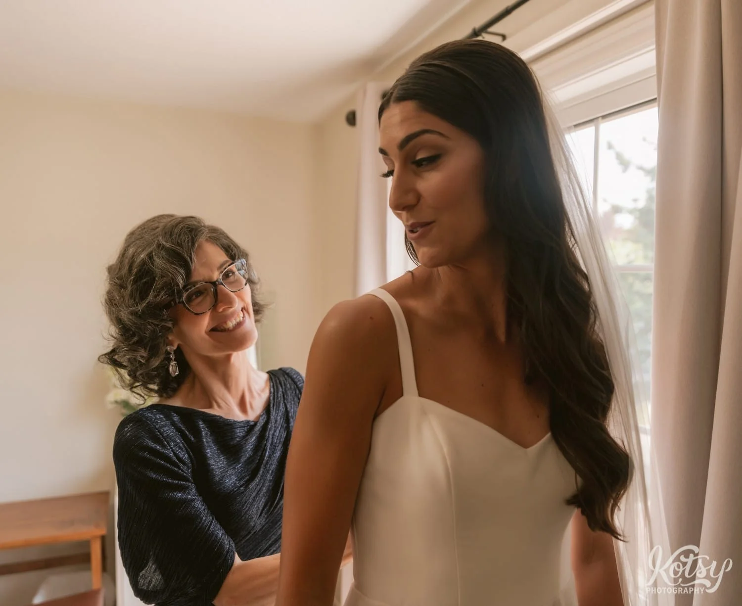 A mother smiles as she zips up her daughter's white wedding gown in a bedroom