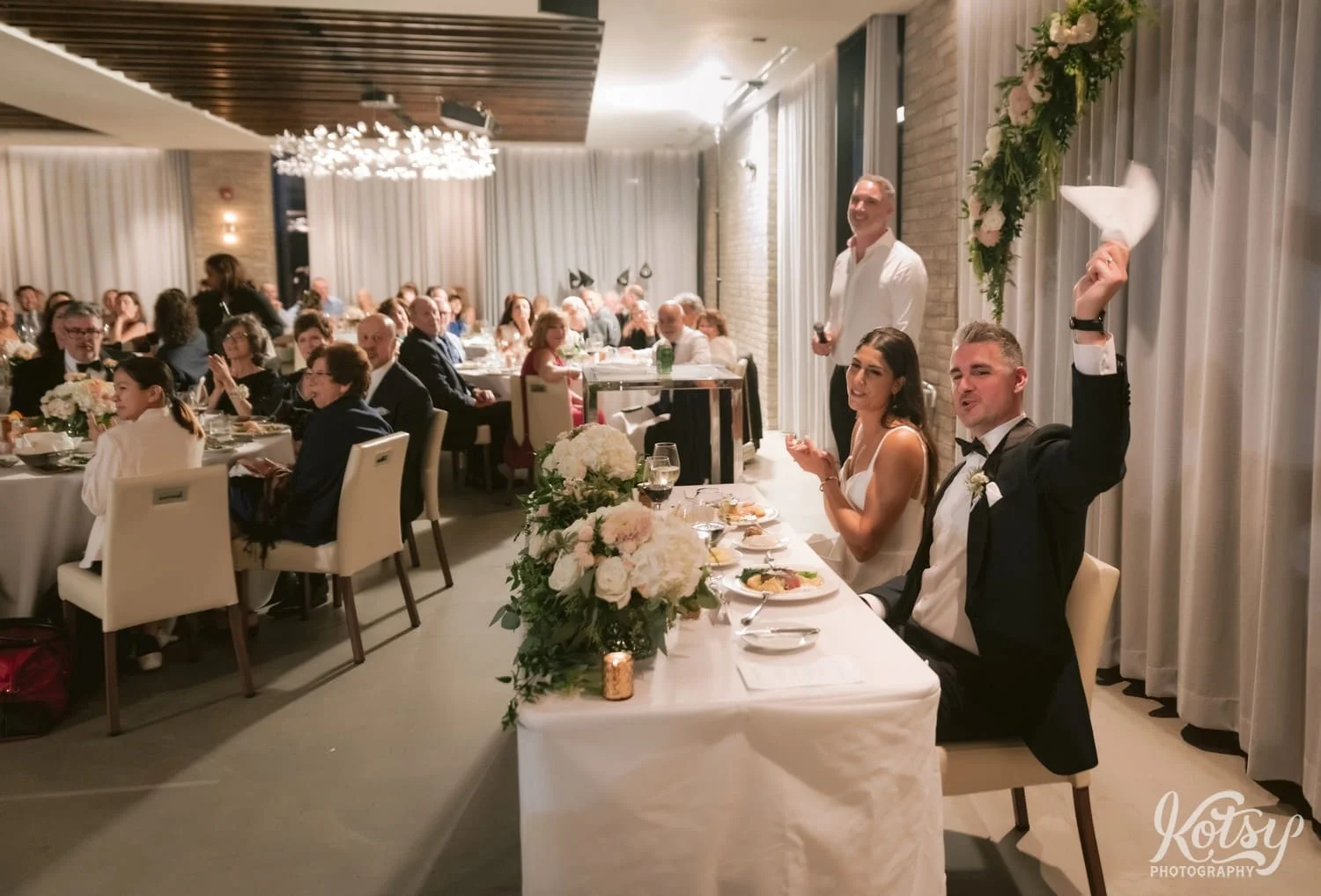 A groom waves a napkin in the area as a room full of people sing happy birthday to his mother at his Village Loft wedding reception in Toronto, Canada