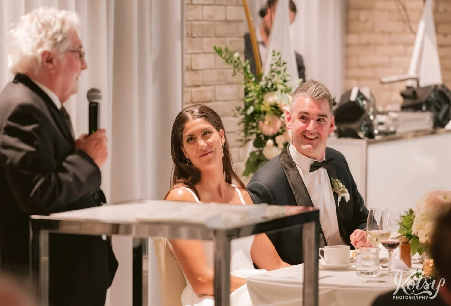 A bride and groom smile as the groom's grandfather makes a speech during a Village Loft wedding reception in Toronto, Canada