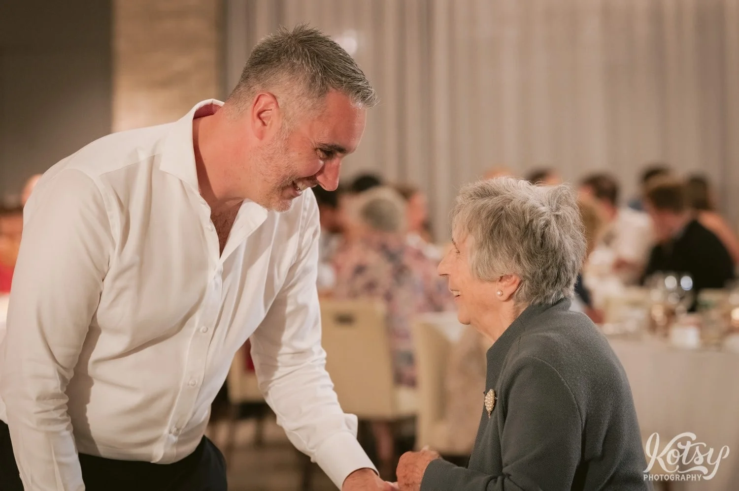 A man smiles as he speaks to his grandmother while holding his hand at a Village Loft wedding reception in Toronto, Canada