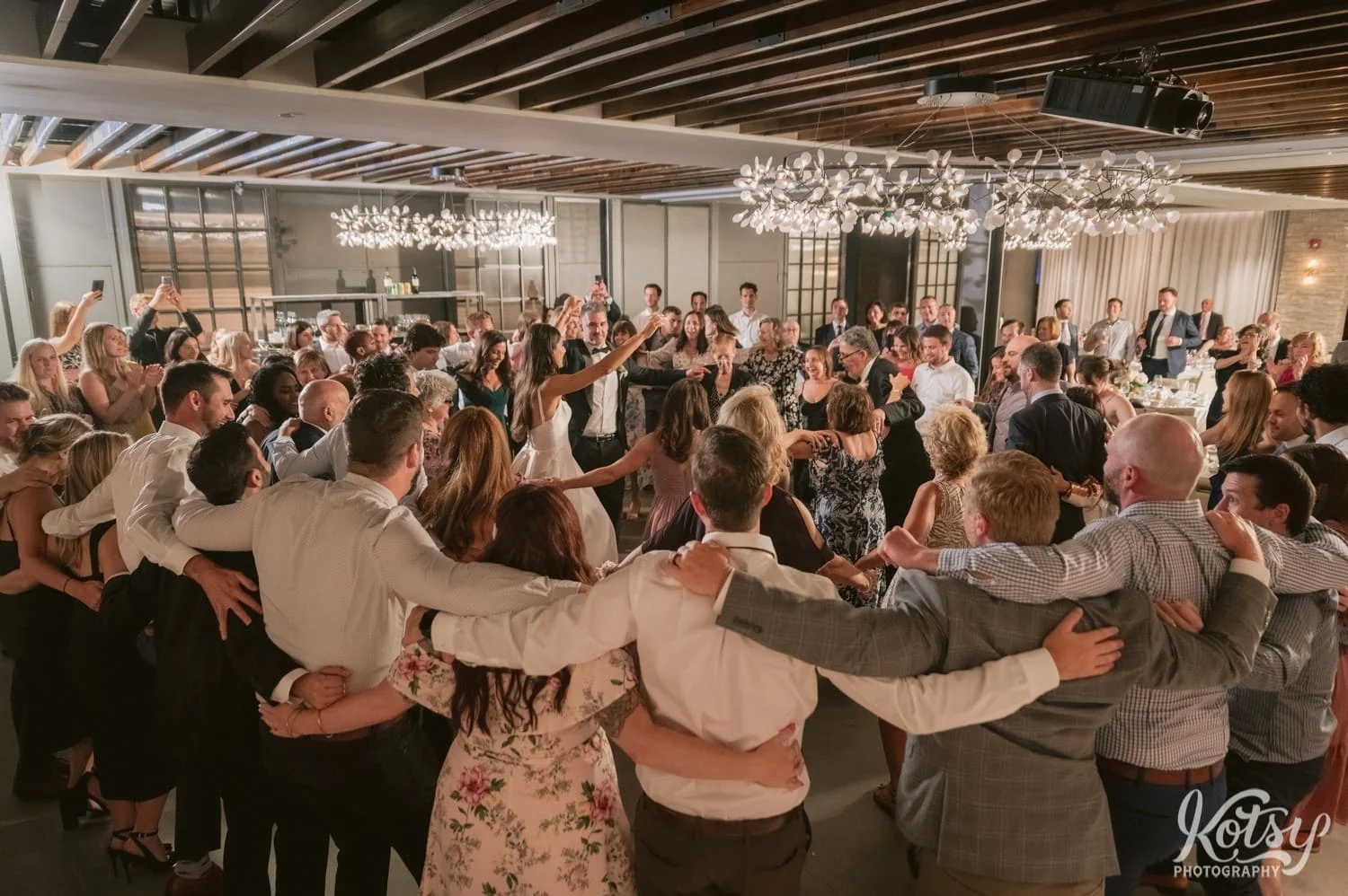 A room full of people dance in a circle with their arms around each other during a Village Loft wedding reception in Toronto, Canada