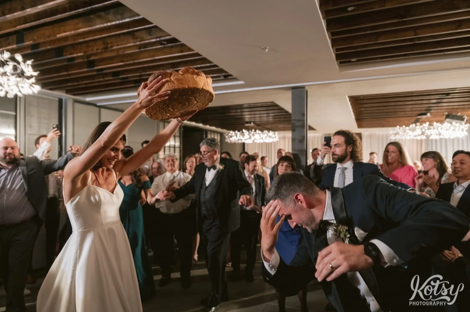 A groom bows at his bride who holds up a large loaf of bread during their Village Loft wedding reception in Toronto, Canada