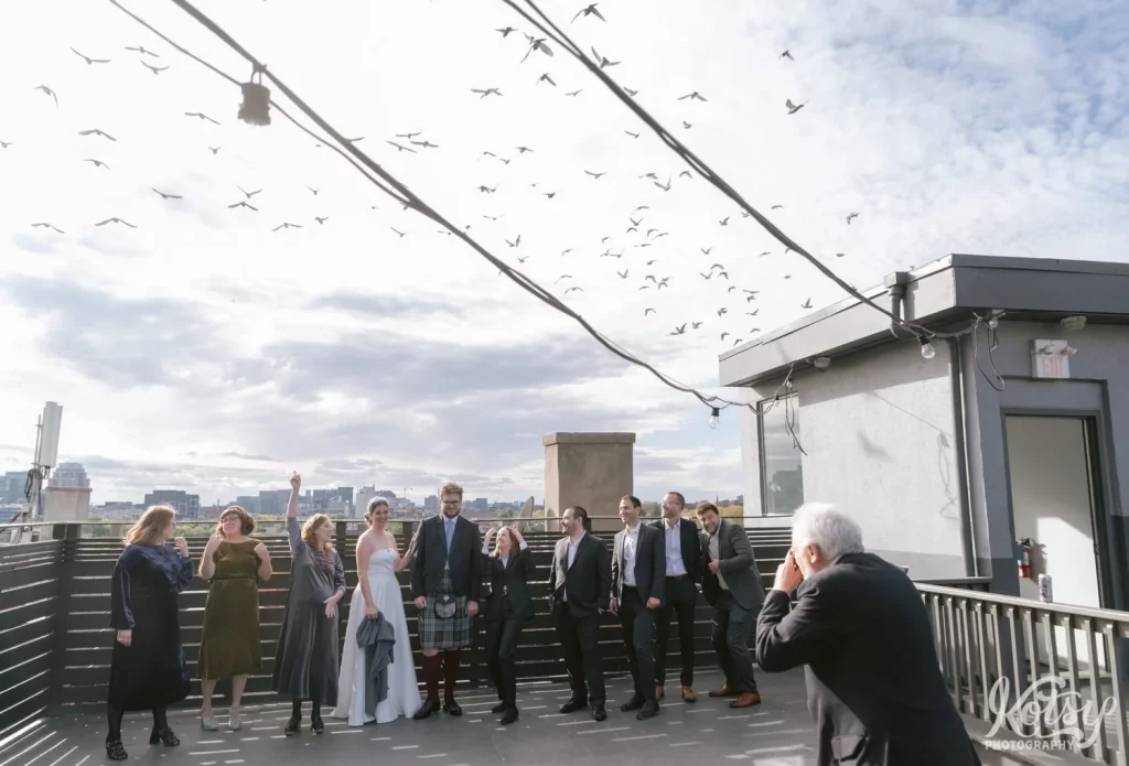 A bright and goom pose with their wedding party as an elderly man takes their photo on a rooftop patio as dozens of seagulls fly overhead. Photographed at The Burroughes Building in Toronto, Canada