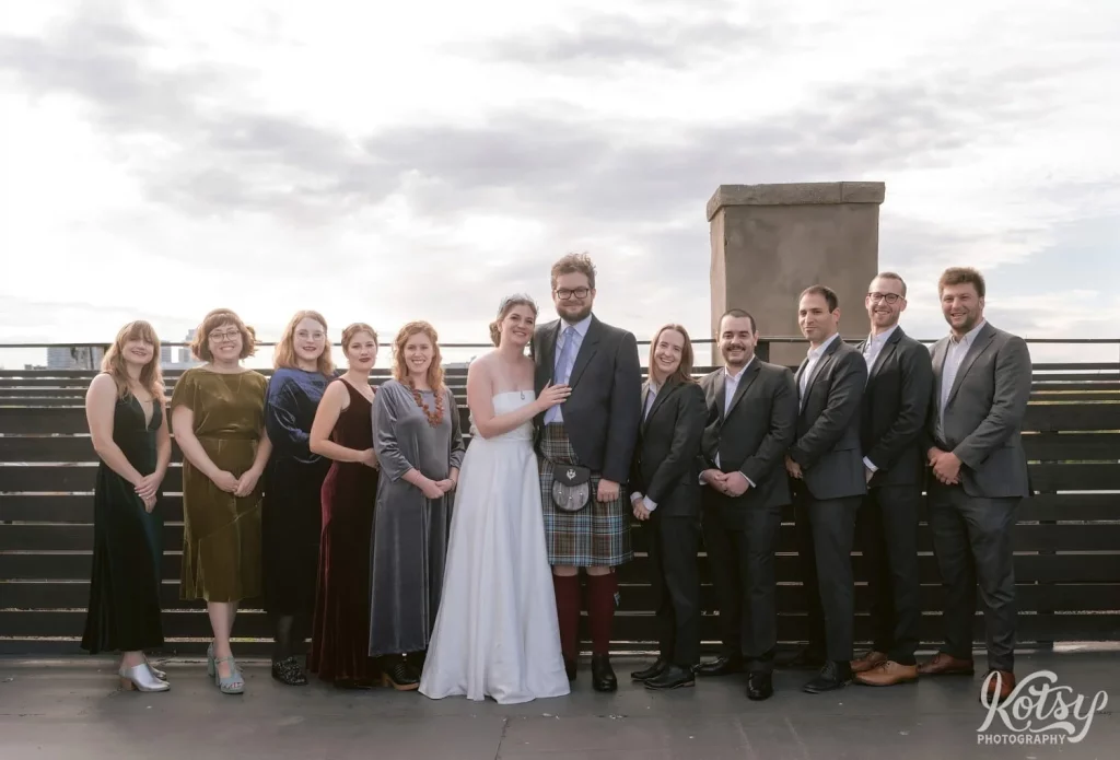 A bride and a white wedding gown and groom wearing a kilt posed with their wedding party on a rooftop patio at The Burroughes Building in Toronto, Canada