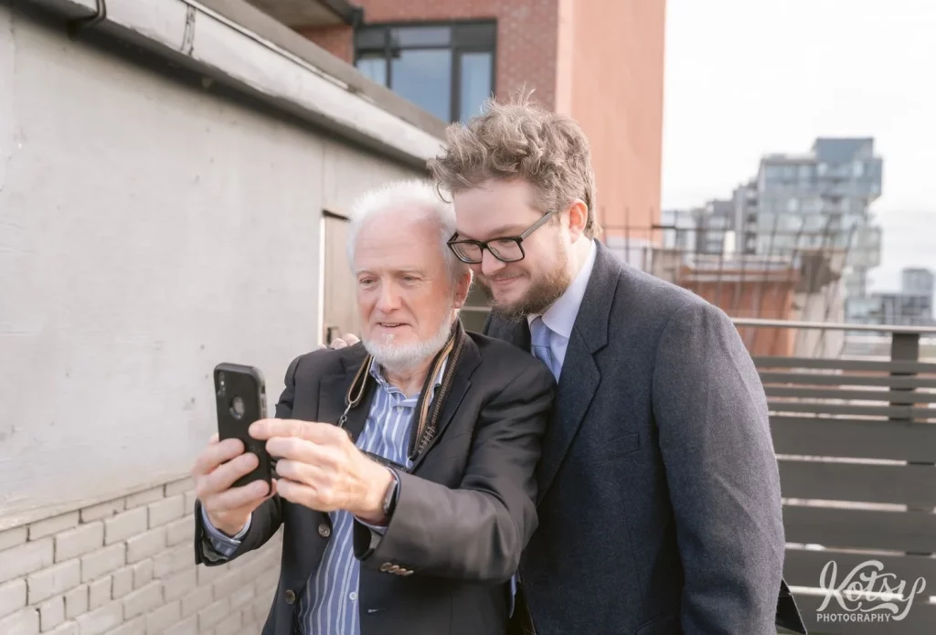 A groom and his father look at a picture on his cell phone on a rooftop patio at The Burroughes Building in Toronto, Canada