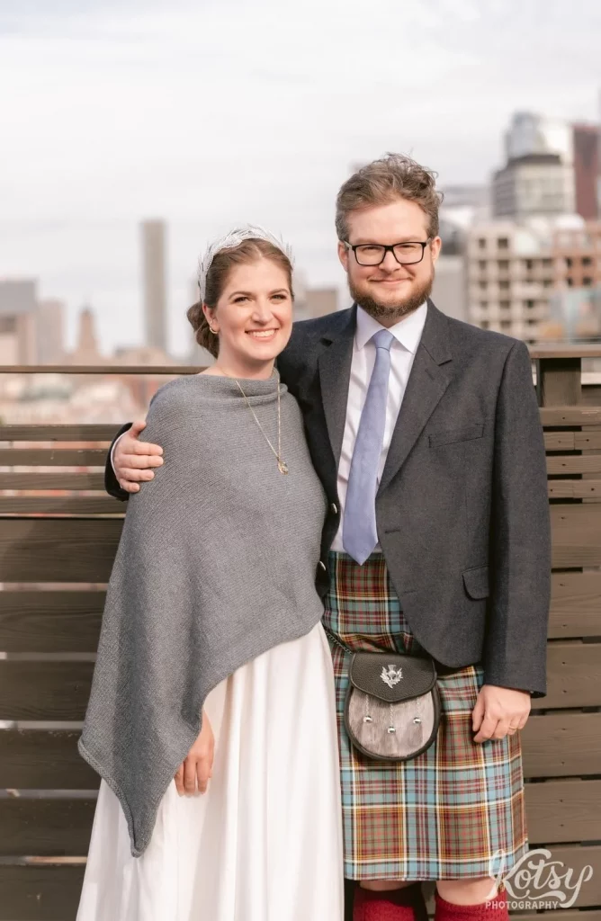A groom wearing a kilt poses for a photo with his arm around his bride wearing a gray shaul over white wedding gown on a rooftop patio at The Burroughes Building in Toronto, Canada