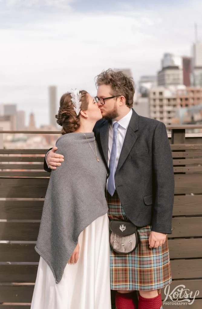A groom wearing a gray blazer and kilt kisses his bride wearing a gray shaul over a white wedding gown on a rooftop patio at The Burroughes Building in Toronto, Canada