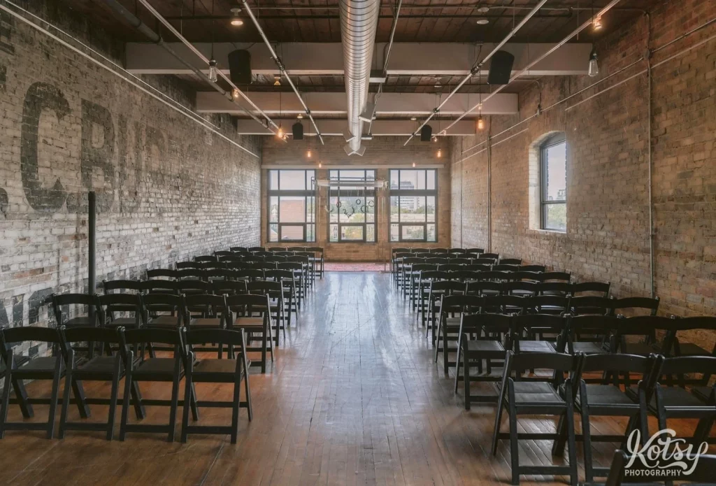 A vacant room with rows of black chairs, brick walls and wooden floors inside the The Burroughes Building