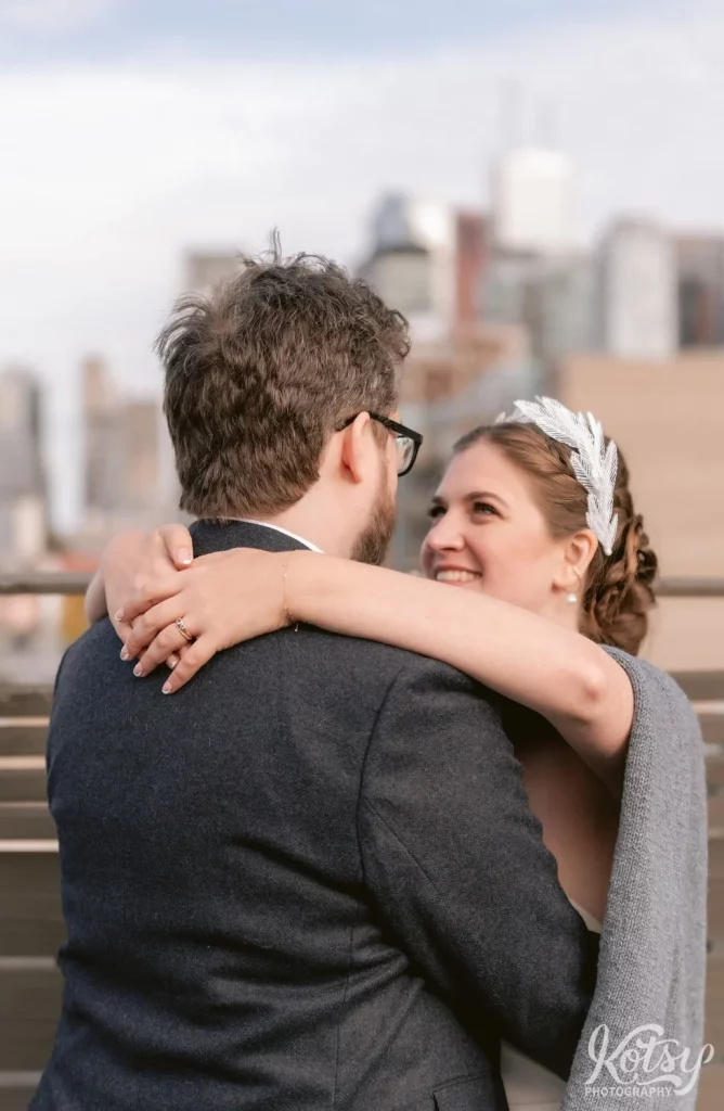 A bride smiles with her arms around her groom's neck as she gazes into his eyes on the rooftop patio during their Burroughes Building wedding in Toronto, Canada