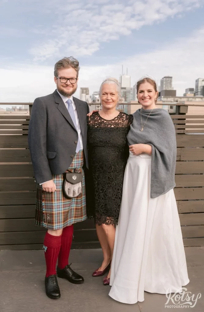 A groom wearing a gray blazer and kilt poses with his mother wearing a black dress and bridewearing a white wedding gown and gray shaul on a rooftop patio during there Burroughes Building wedding in Toronto, Canada