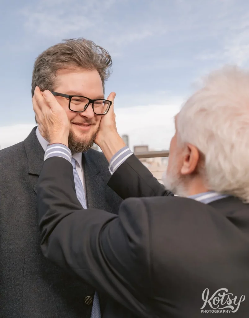 A man in glasses and a beard smiles as his father places his hands on his cheeks with a blue sky behind him.