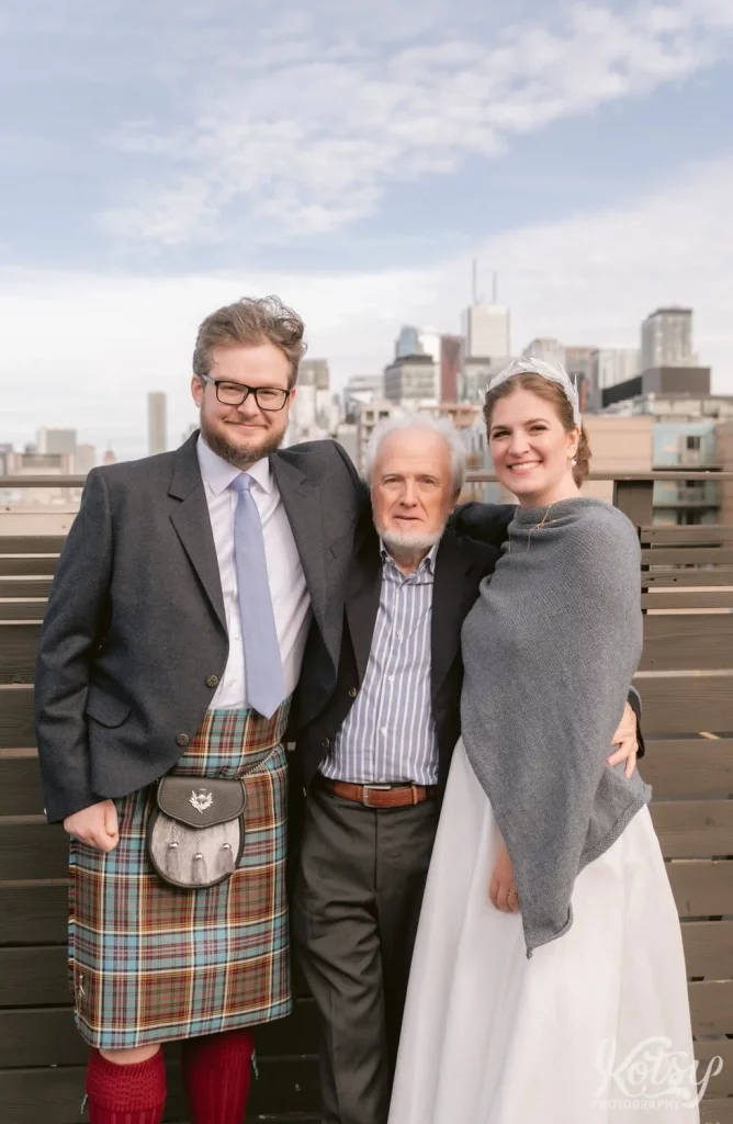A groom wearing a kilt and bride wearing a gray shaul over white wedding gown posed with his father on a rooftop patio with the Toronto skyline in the background