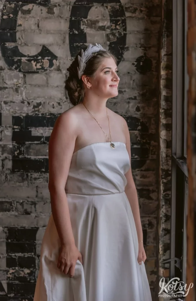 A bride in a white wedding gown, gold necklace and white tiara stands in front of a gray brick wall looking out a window during her Burroughes Building wedding in Toronto, Canada