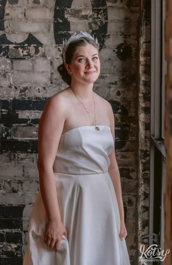 A bride wearing a white wedding gown, gold necklace and white tiara stands in front of a gray brick wall and smiles for the camera during her Burroughes Building wedding in Toronto, Canada