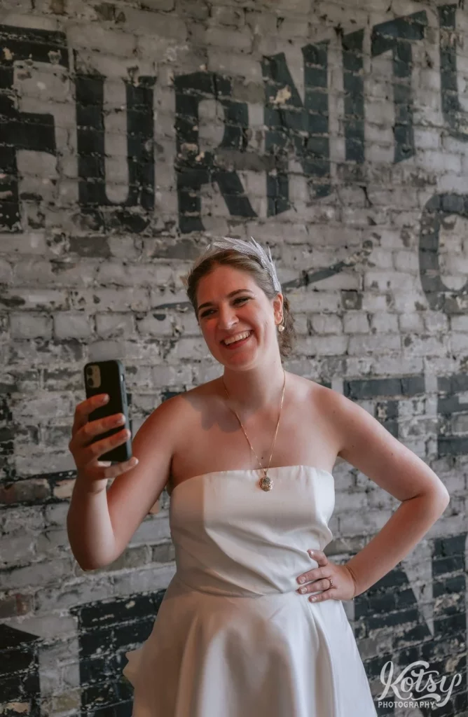 A bride wearing a white wedding gown smiles as she looks at her phone in front of a gray wall during her Burroughes Building wedding in Toronto, Canada