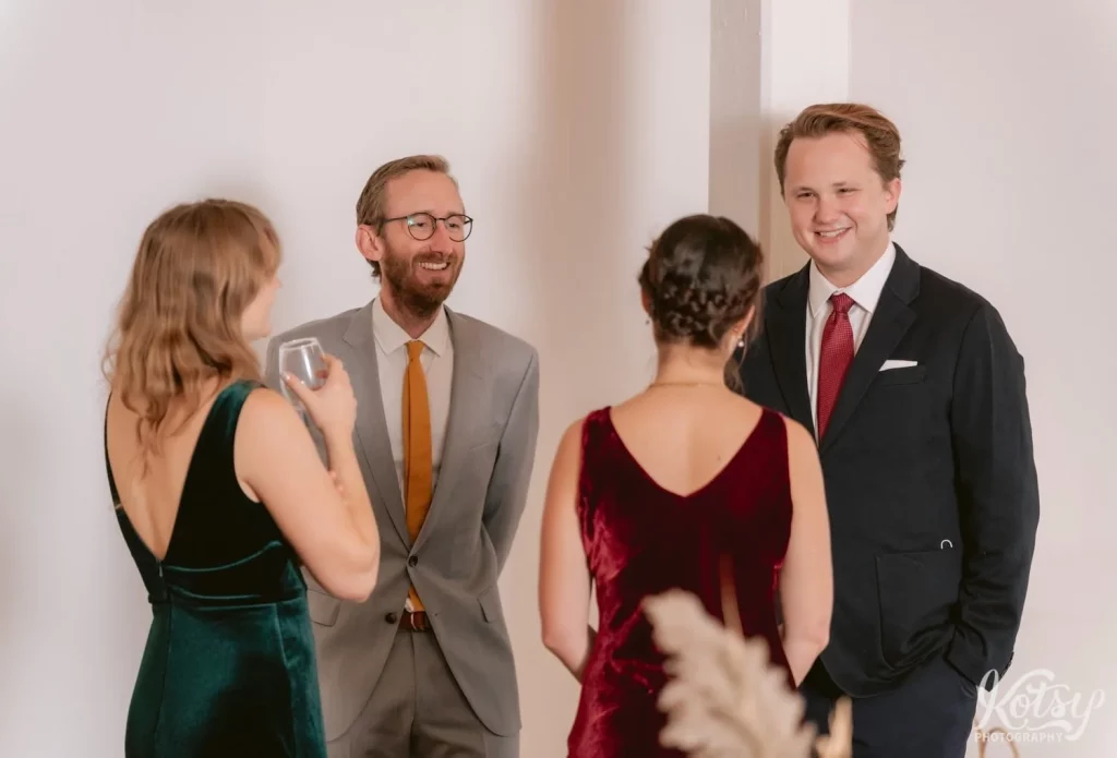 Two men in suits smile as they talk to women in the foreground facing them during Burroughes Building wedding in Toronto, Canada