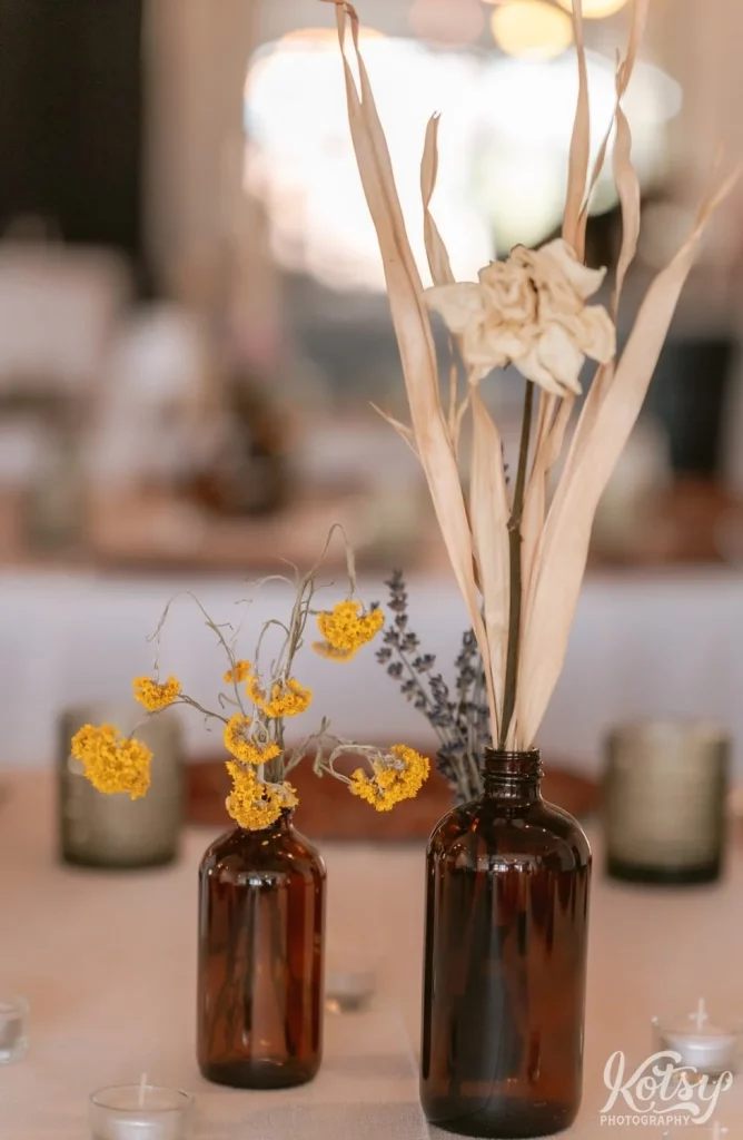 Dried marigolds and dried white rose in brown bottles on a table with white tablecloth.