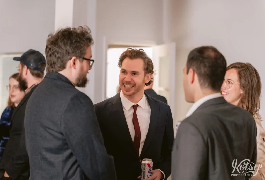 A man in a dark blue suit and red tie smiles as he talks to a man facing him in the foreground during a Burroughes Building wedding in Toronto, Canada
