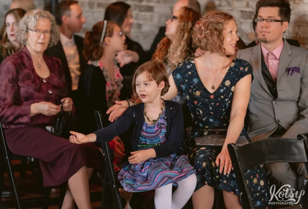 A young girl wearing a blue and purple dress smiles to someone off camera as she sits in a chair amongst a big group of people during a Burroughes Building wedding in Toronto, Canada