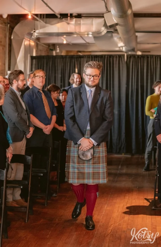 A groom wearing glasses, gray blazer, kilt and red stockings walks down the aisle during his Burroughes Building wedding ceremony in Toronto, Canada