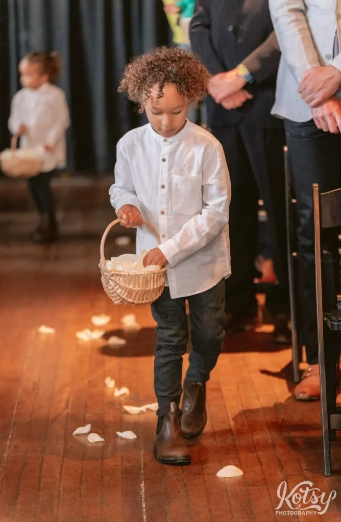 A child and a white dress shirt walks down the aisle with his hand inside a basket of white rose petals during a Burroughes Building wedding ceremony in Toronto, Canada