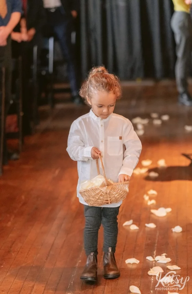 A young girl wearing a white dress shirt walks down an aisle with her hand in a basket holding white rose petals during a Burroughes Building wedding ceremony in Toronto, Canada