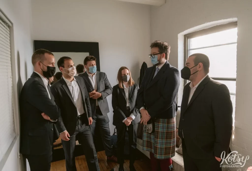 A group of men in dark suits and a groom in a kilt stand in a room with masks on.