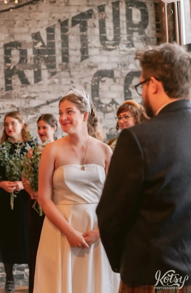 A bride wearing a white wedding gown smiles to someone off camera as she stands at the altar during her Burroughes Building wedding ceremony in Toronto, Canada