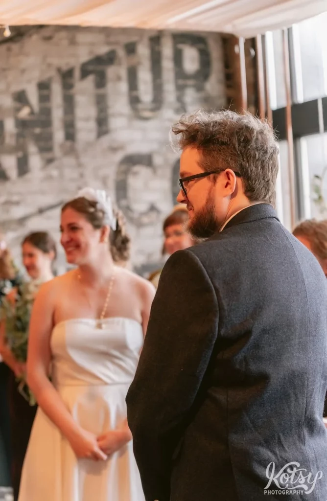 A groom wearing a gray blazer glasses and a beard smiles to someone off camera while at the altar during his Burroughes Building wedding ceremony in Toronto, Canada