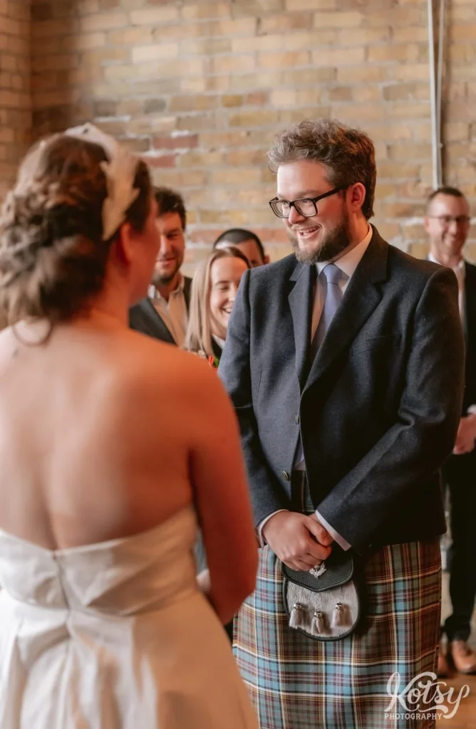 A groom wearing a gray blazer blue tie and kilt smiles as he looks at the officiant during his Burroughes Building wedding ceremony in Toronto, Canada