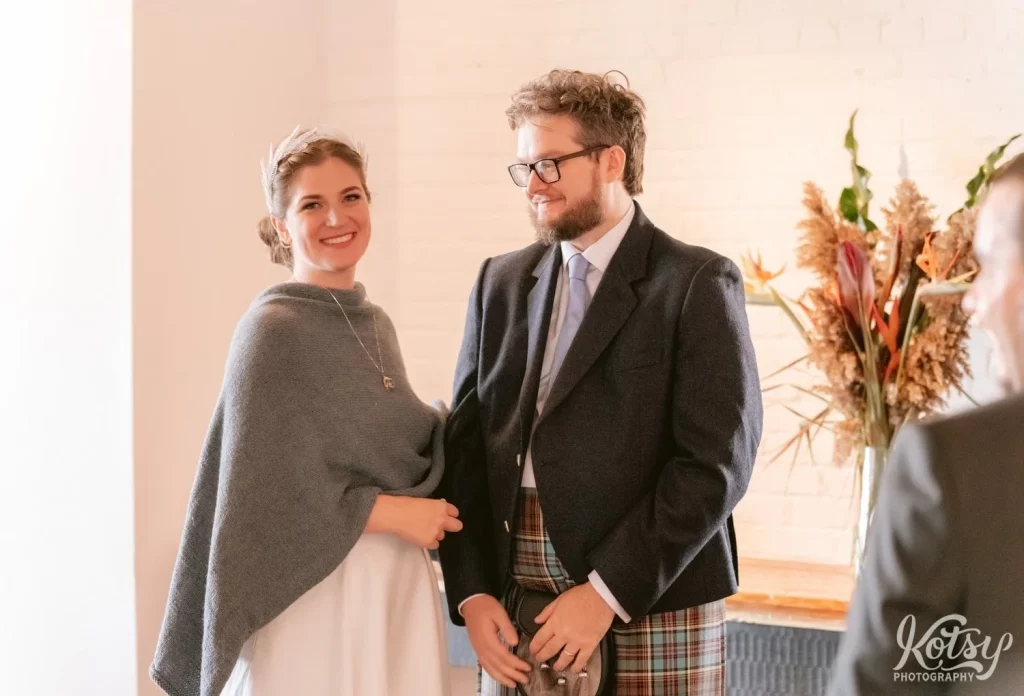 A bride wearing a white wedding gown and gray shaul stan next to her groom wearing a dark gray blazer and kilt