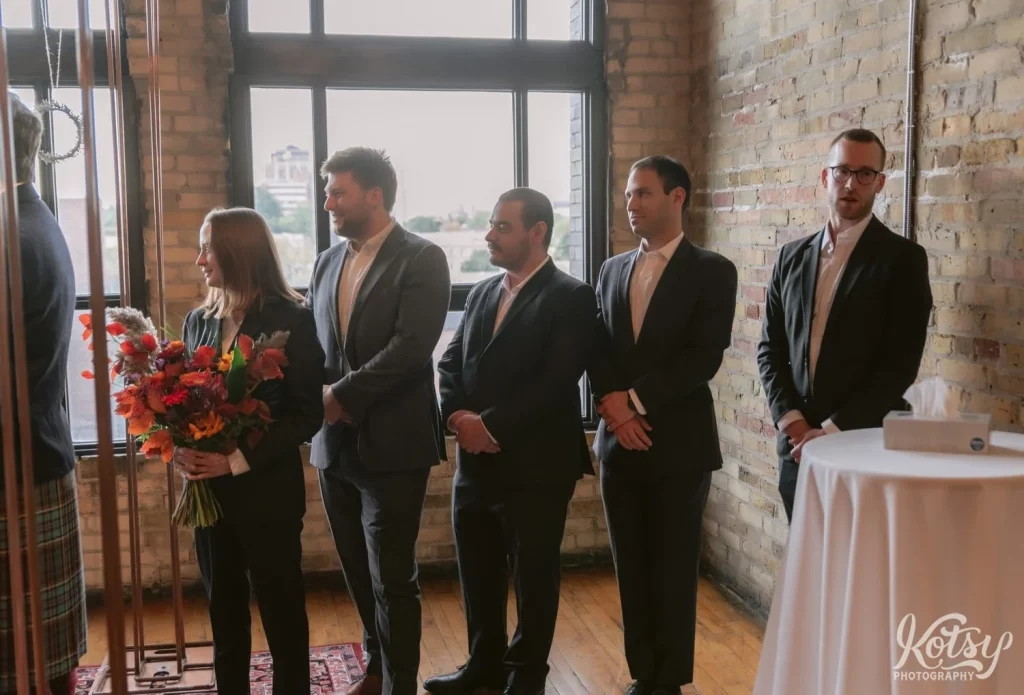 5 groomsmen in black suits look on at their friend during his Burroughes Building wedding ceremony in Toronto, Canada